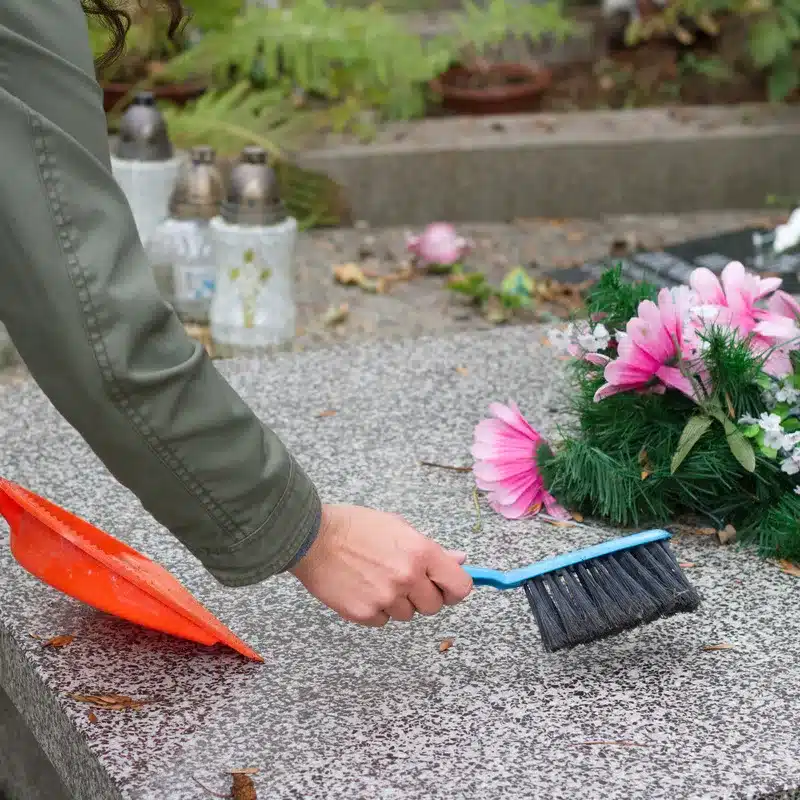 Personne entretenant une pierre tombale avec une brosse, enlevant les feuilles et la poussière pour préserver la propreté du monument funéraire.