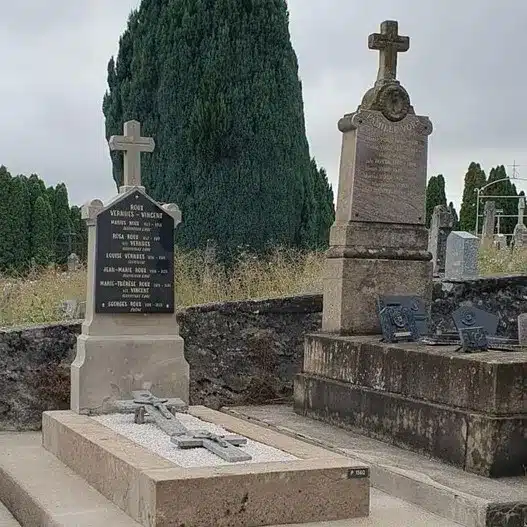Tombe avec stèle en pierre gravée et croix, située dans un cimetière ancien, entourée d’autres sépultures traditionnelles.